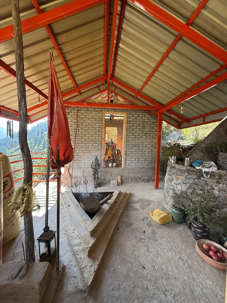 Inside the Manali Ashram: the sacred fire pit beneath the red-trussed roof, with the Devi enshrined beyond